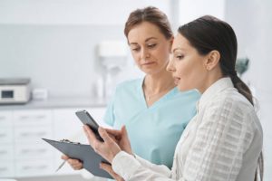 Female person pointing at smartphone display while discussing treatment with beautician in cosmetology clinic